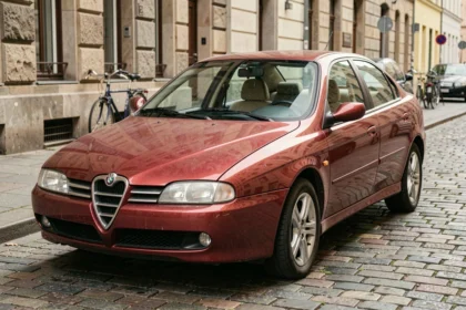 Alfa Romeo 147 youngtimer rouge métallique garée sur des pavés dans une rue européenne, vue 3/4 avant